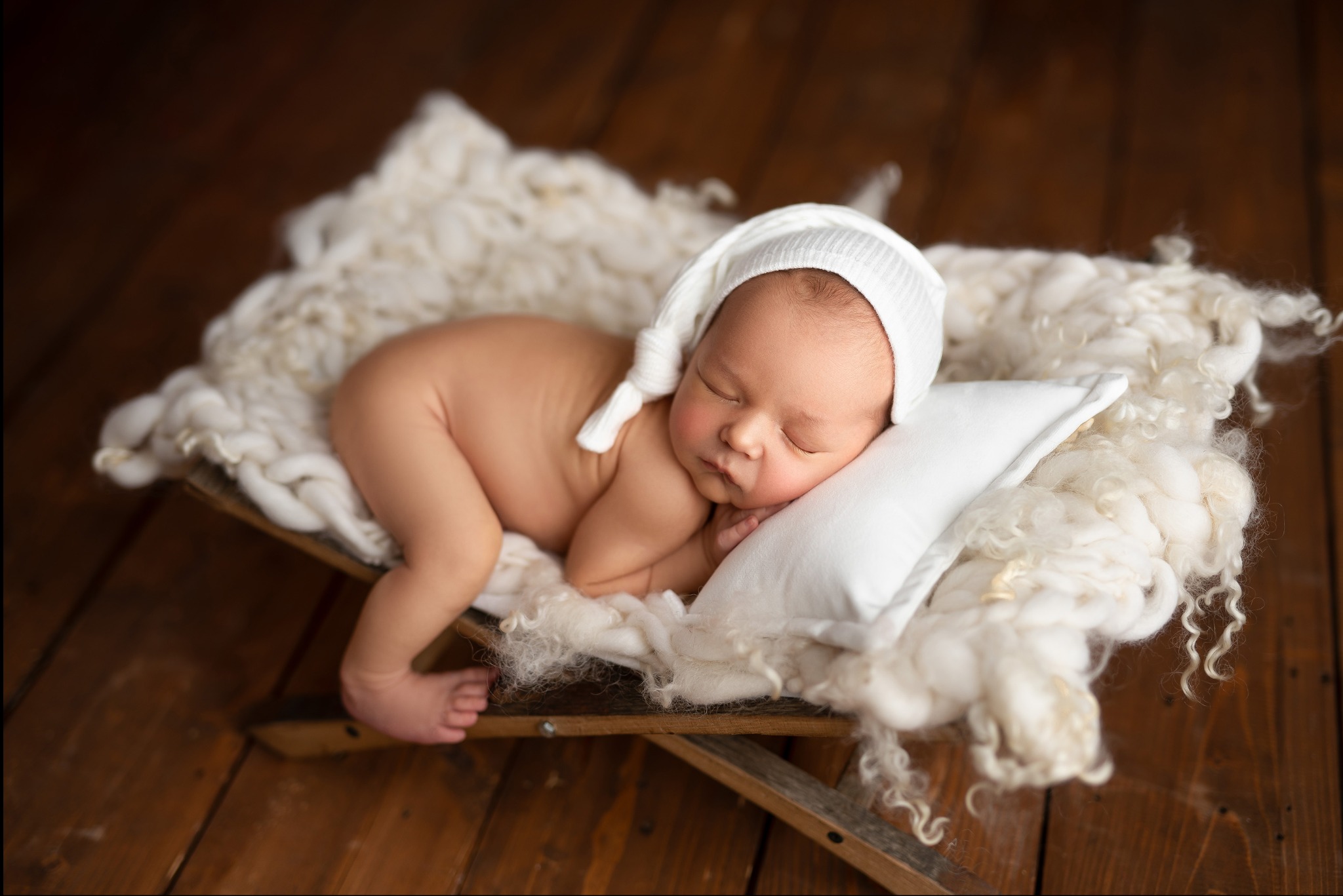 Śpiący noworodek w białej czapeczce, ułożony na puszystym, kremowym kocyku i białej poduszce w drewnianym, niskim łóżeczku na tle drewnianej podłogi.