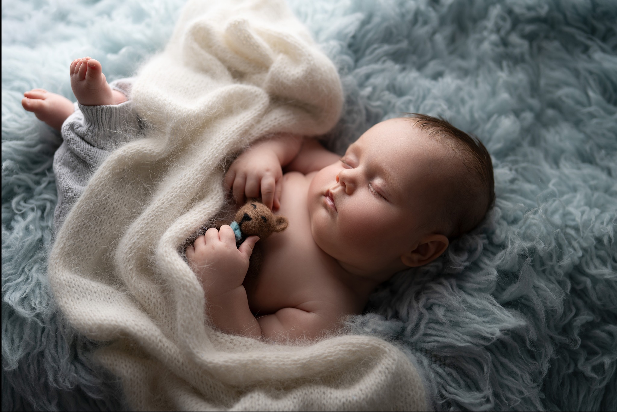 Śpiący noworodek owinięty w kremowy koc na teksturowanym, błękitnym tle, trzymający małego pluszowego misia.