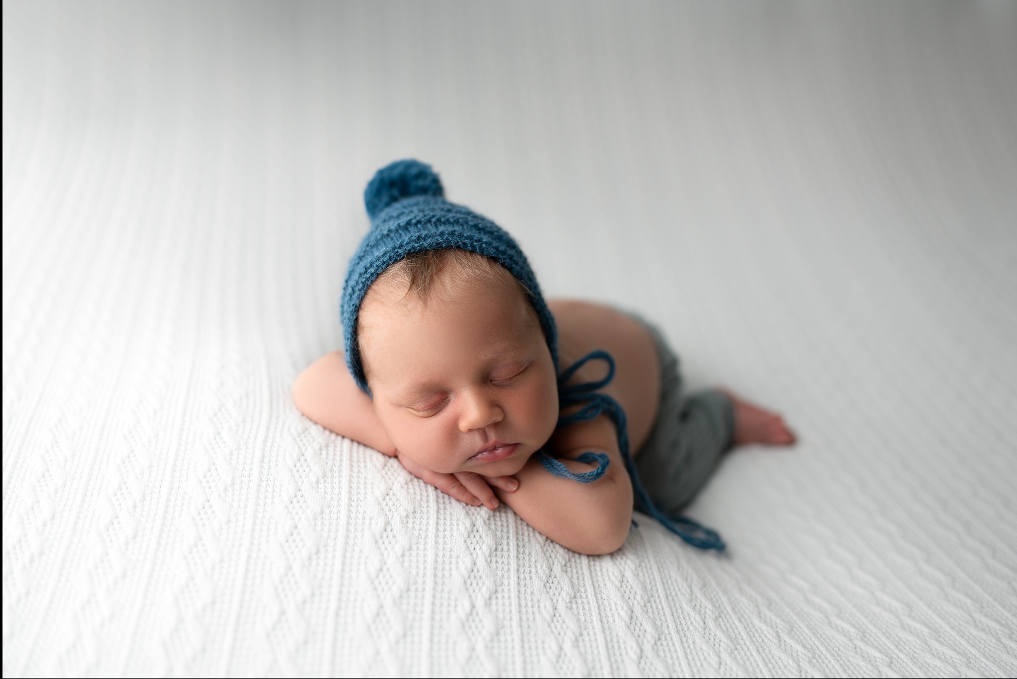 Śpiący noworodek w niebieskiej, dzierganej czapeczce z pomponem, leżący na białym, teksturowanym kocyku.