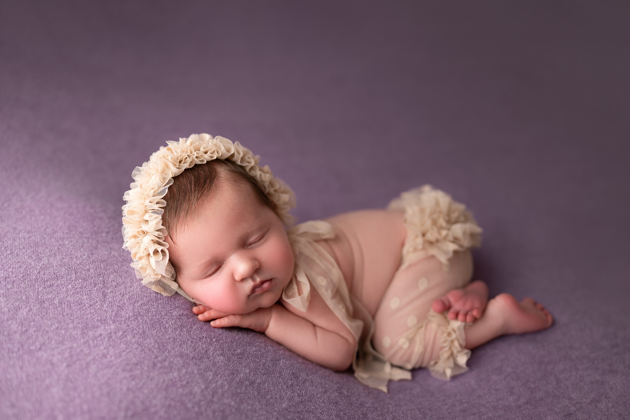 Śpiące niemowlę w beżowym, koronkowym czepku i spodenkach z falbankami na fioletowym tle.