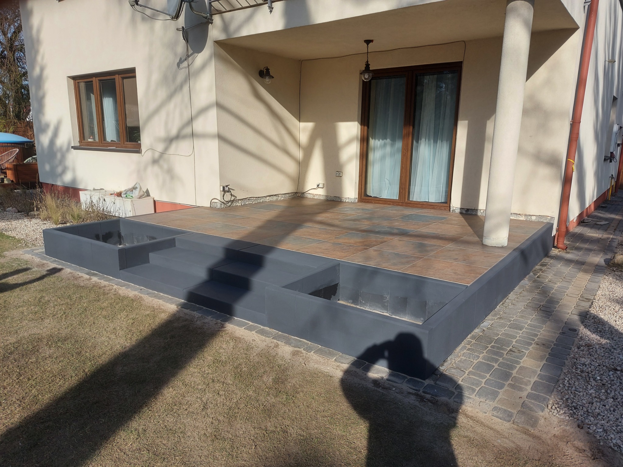 Wykończony taras z brązowymi płytkami, szare schody z obrzeżem z kostki brukowej, widok z zewnątrz w słoneczny dzień.