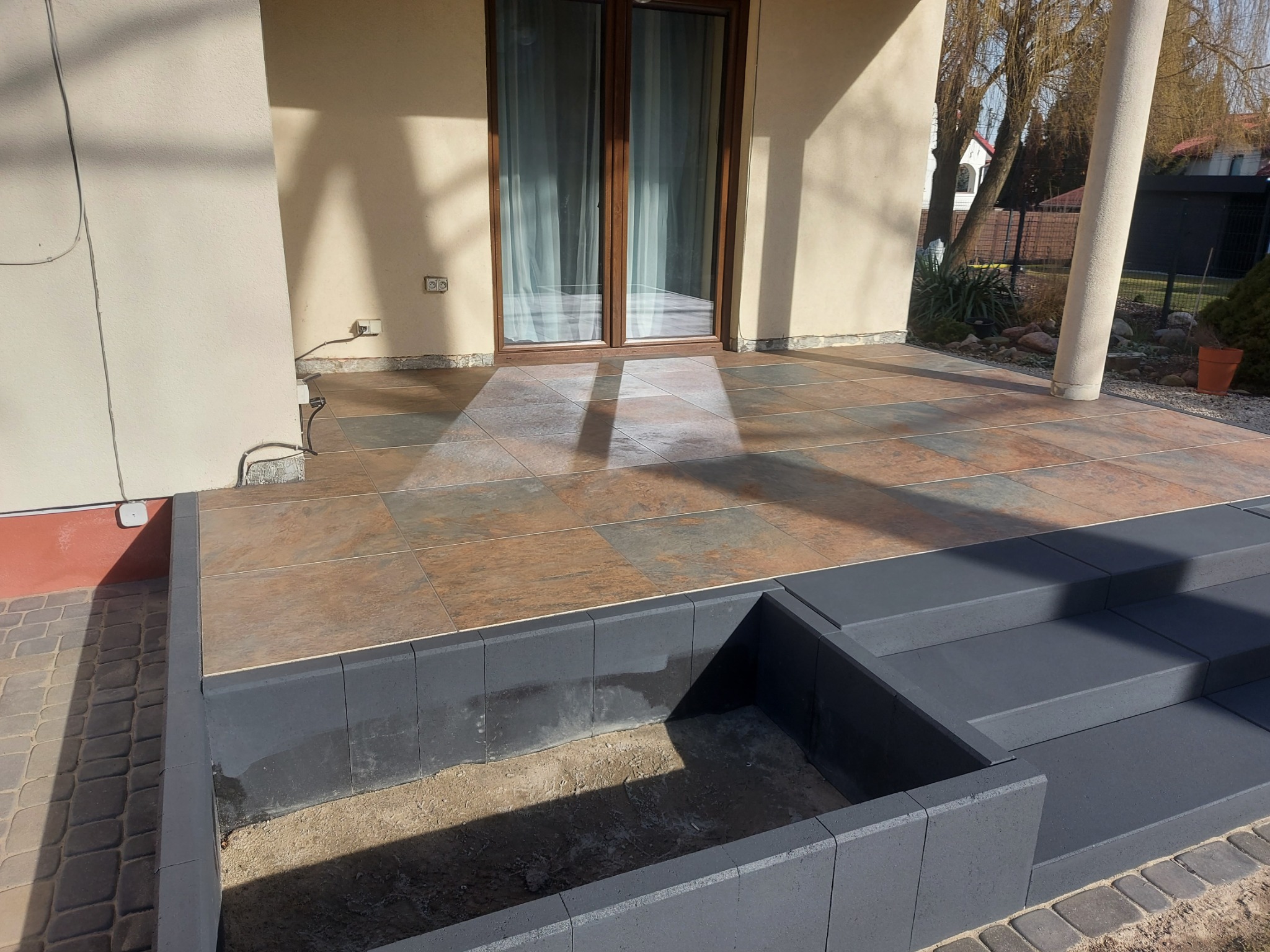 Wykończony taras z brązowych płytek z szarą obudową schodów i rabaty kwiatowej w trakcie budowy, widok na dwuskrzydłowe drzwi balkonowe i fragment ogrodu.