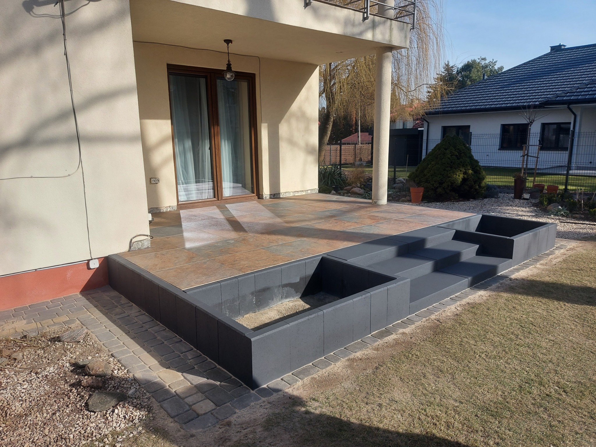 Wykończony taras z brązowych płyt ceramicznych, zintegrowany z betonowymi schodami i obrzeżem z kostki, widok na dom z ogrodem w słoneczny dzień.