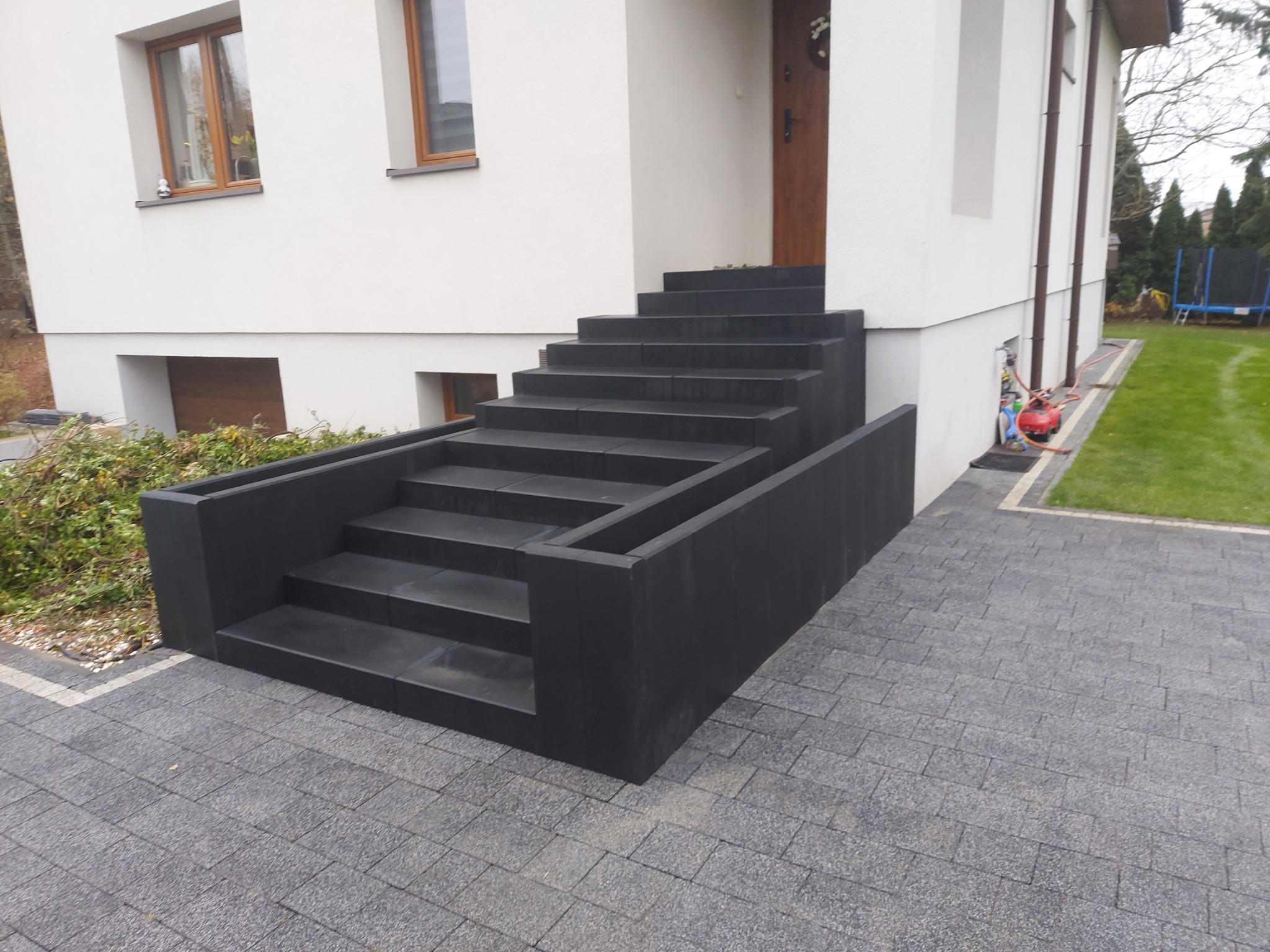 Nowoczesne, czarne schody wejściowe z kostki brukowej prowadzące do białego domu, otoczone szarą kostką brukową i zielonym trawnikiem.