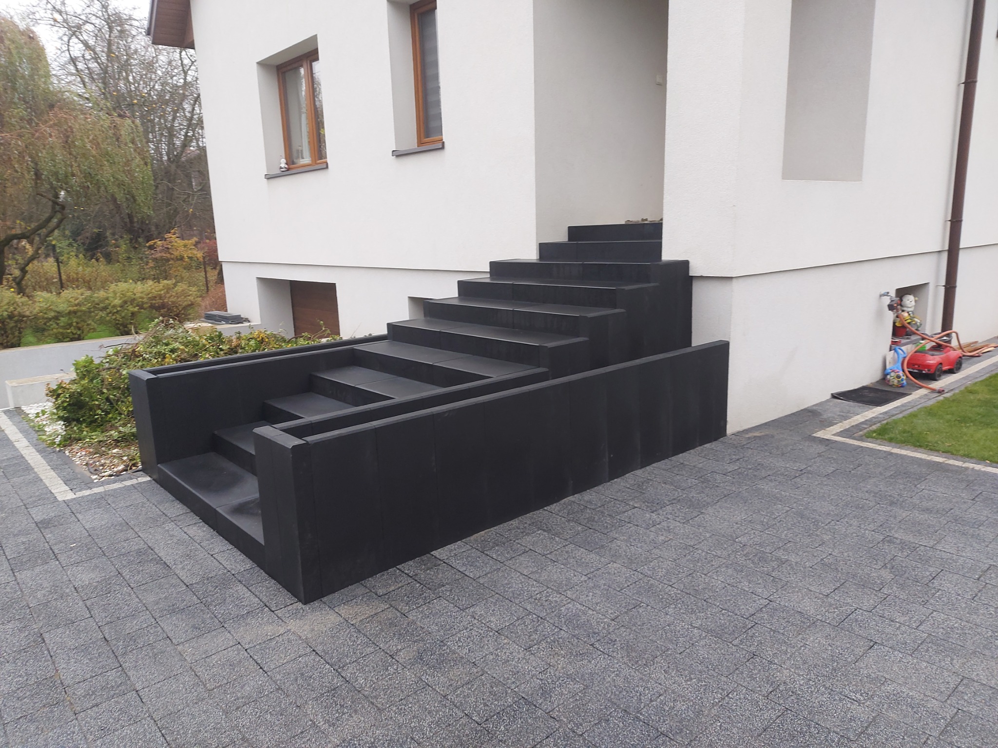 Nowoczesne, ciemnoszare schody wejściowe z kostki brukowej prowadzące do białego domu z szarą kostką brukową na podjeździe. Widoczny fragment trawnika i roślinność w tle.