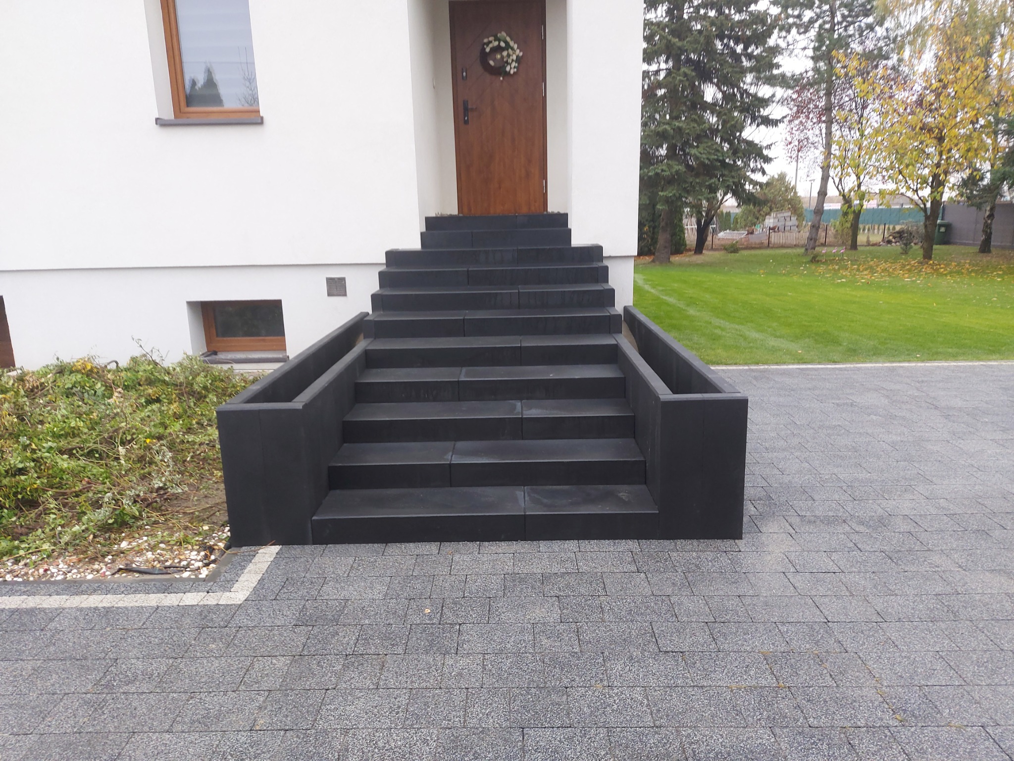 Nowoczesne schody wejściowe z czarnej kostki prowadzące do drzwi z wieńcem, otoczone szarą kostką brukową na podjeździe.