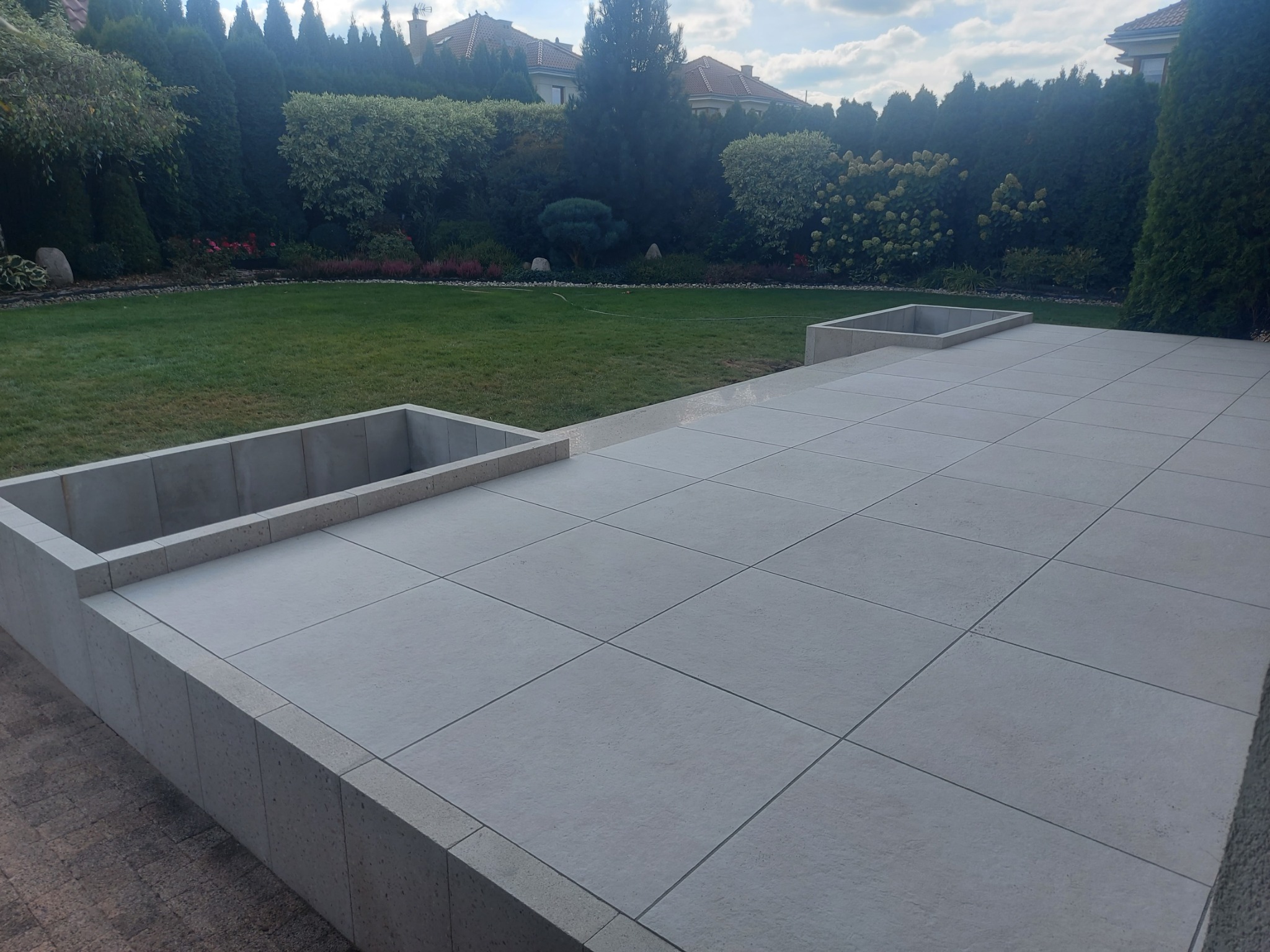 Nowoczesny taras z jasnoszarych płyt betonowych w ogrodzie z zielonym trawnikiem i ozdobnymi krzewami, widoczne betonowe obrzeża i donice.