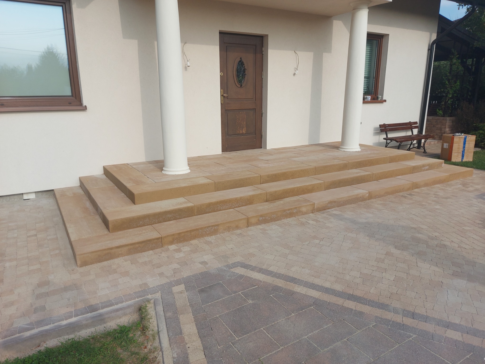 Wejście do domu z kolumnami i schodami z beżowej kostki, otoczone chodnikiem z kostki w dwóch odcieniach. Widoczna drewniana ławka i paczka w tle.