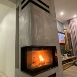 Nowoczesny kominek obudowany szarym betonem architektonicznym z poziomymi czarnymi wstawkami, płomienie widoczne przez szybę.