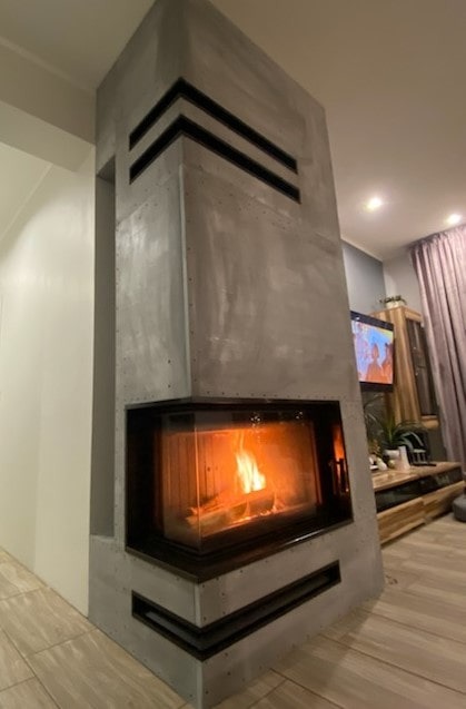 Nowoczesny kominek obudowany szarym betonem architektonicznym z poziomymi czarnymi wstawkami, płomienie widoczne przez szybę.