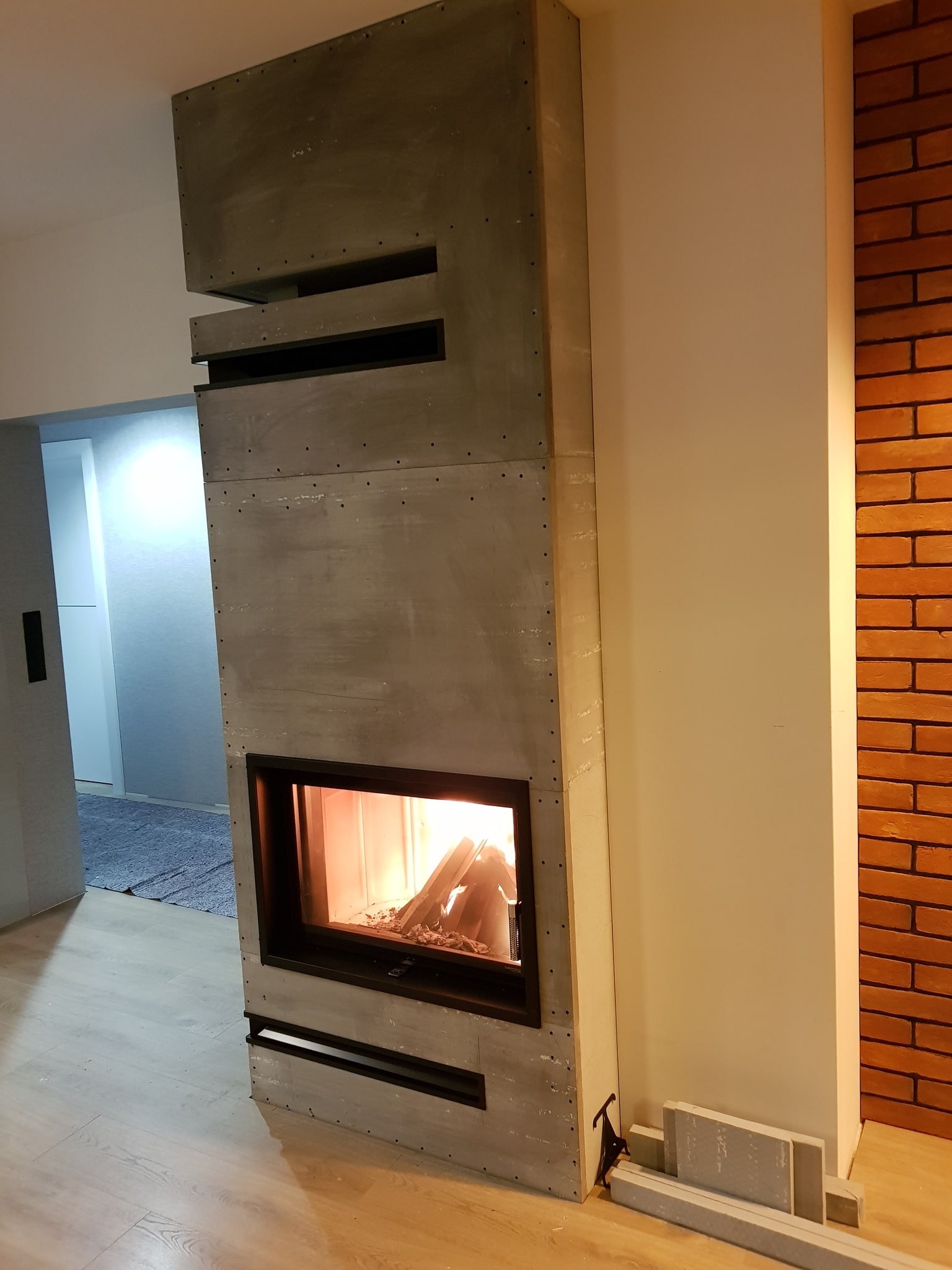 Nowoczesny kominek z obudową z szarego betonu architektonicznego, z widocznym paleniskiem i płomieniami, w trakcie montażu, z fragmentem ściany z cegły w tle.