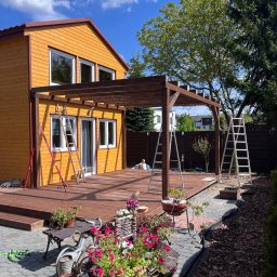 Vadym Boiko - Żółty dom z drewnianą pergolą w trakcie montażu, widoczne drabiny i kwiaty w donicach. Ujęcie w słoneczny dzień z niebieskim niebem.