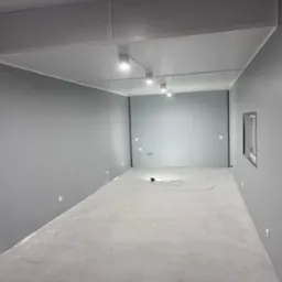 Szare, puste wnętrze po remoncie z nowym oświetleniem punktowym na suficie podwieszanym i widocznymi gniazdkami elektrycznymi na ścianach.