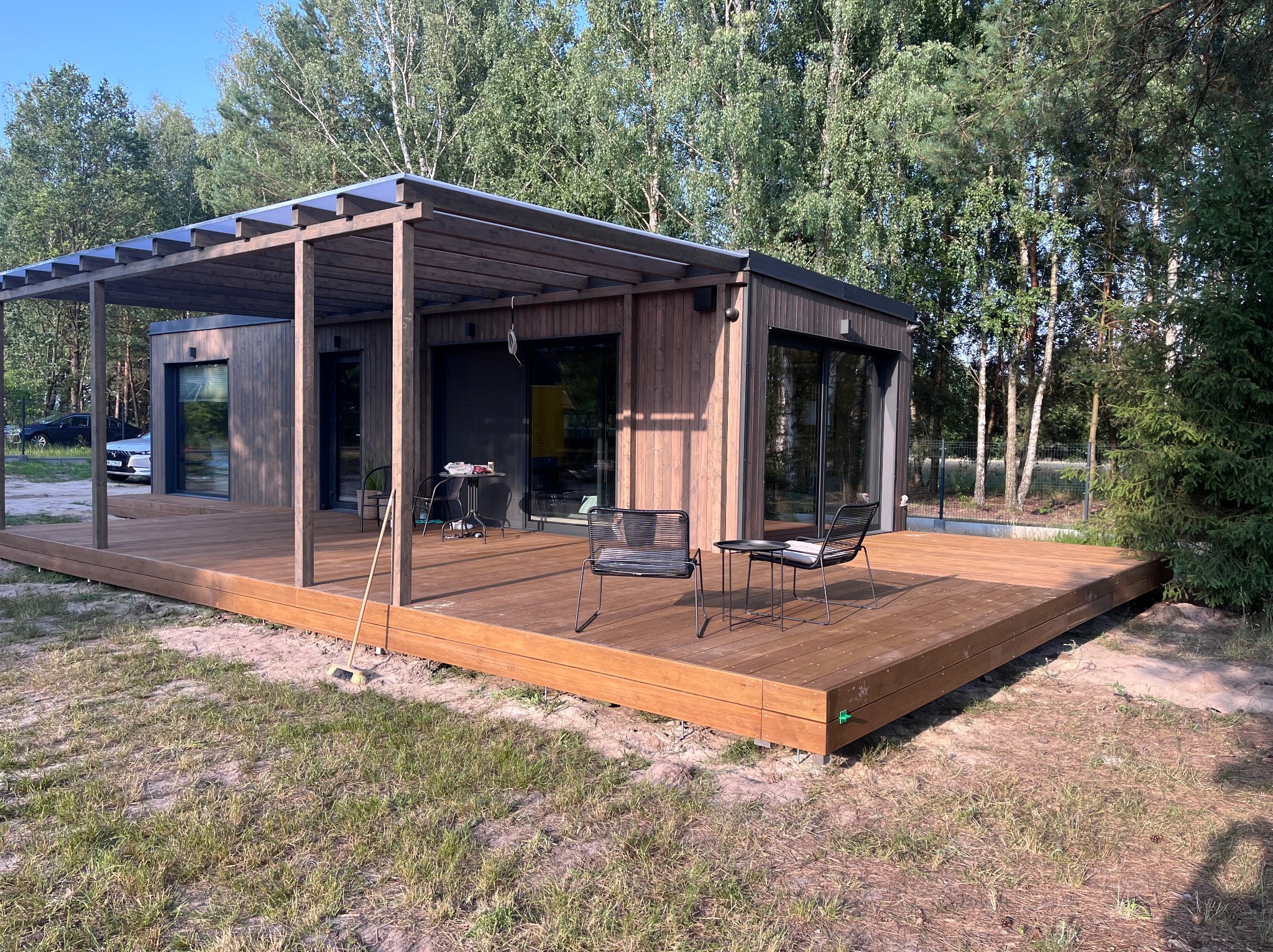 Nowoczesna, drewniana altana z zadaszeniem i tarasem w otoczeniu zieleni. Widoczne krzesła ogrodowe i starannie wykończona podłoga tarasu.