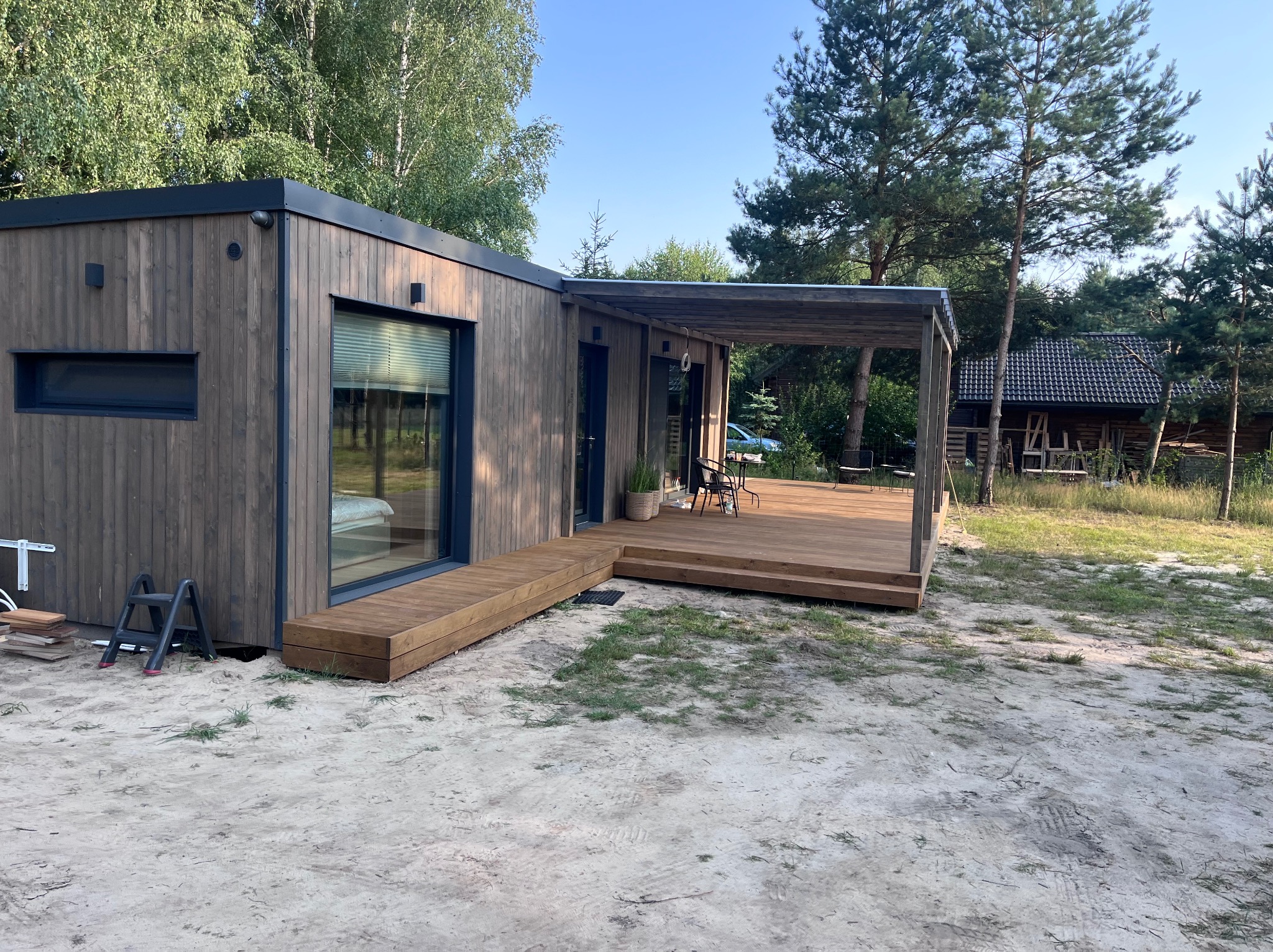 Nowoczesny, drewniany dom modułowy z tarasem i pergolą w naturalnym otoczeniu. Widoczne detale wykończenia i elementy krajobrazu, drabina oparta o ścianę budynku.