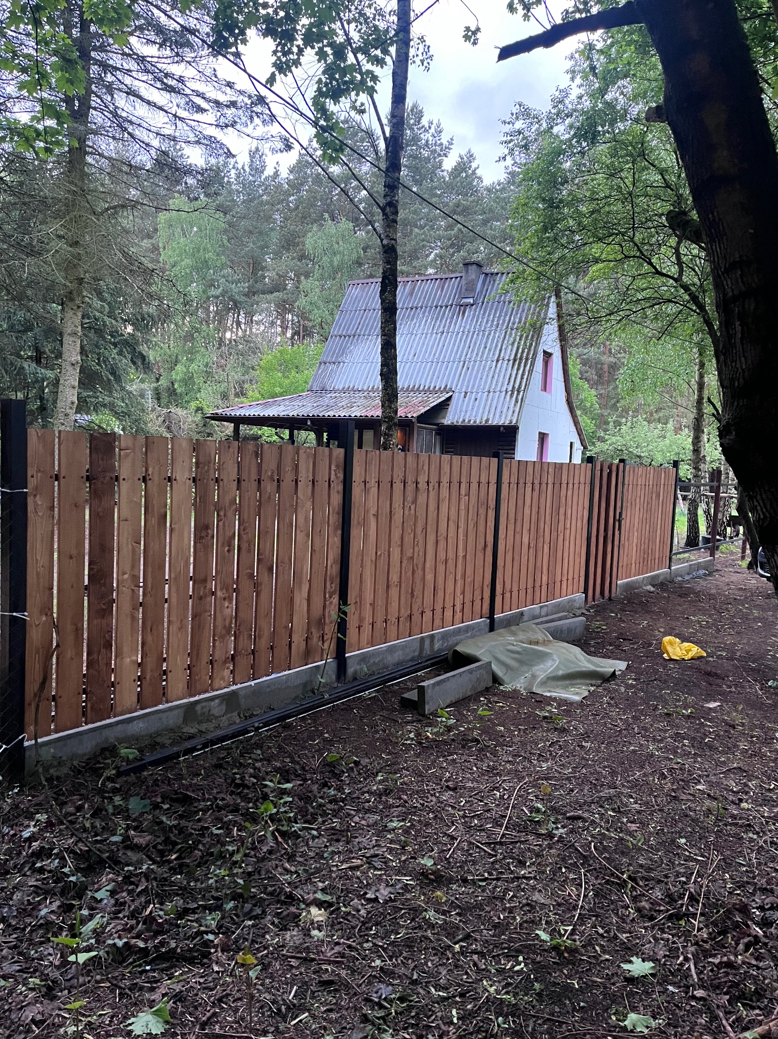 Nowy, drewniany płot panelowy z czarnymi słupkami na tle leśnej działki w Warszawie. Widoczny fragment domu z szarym dachem. Świeżo wykonana instalacja.