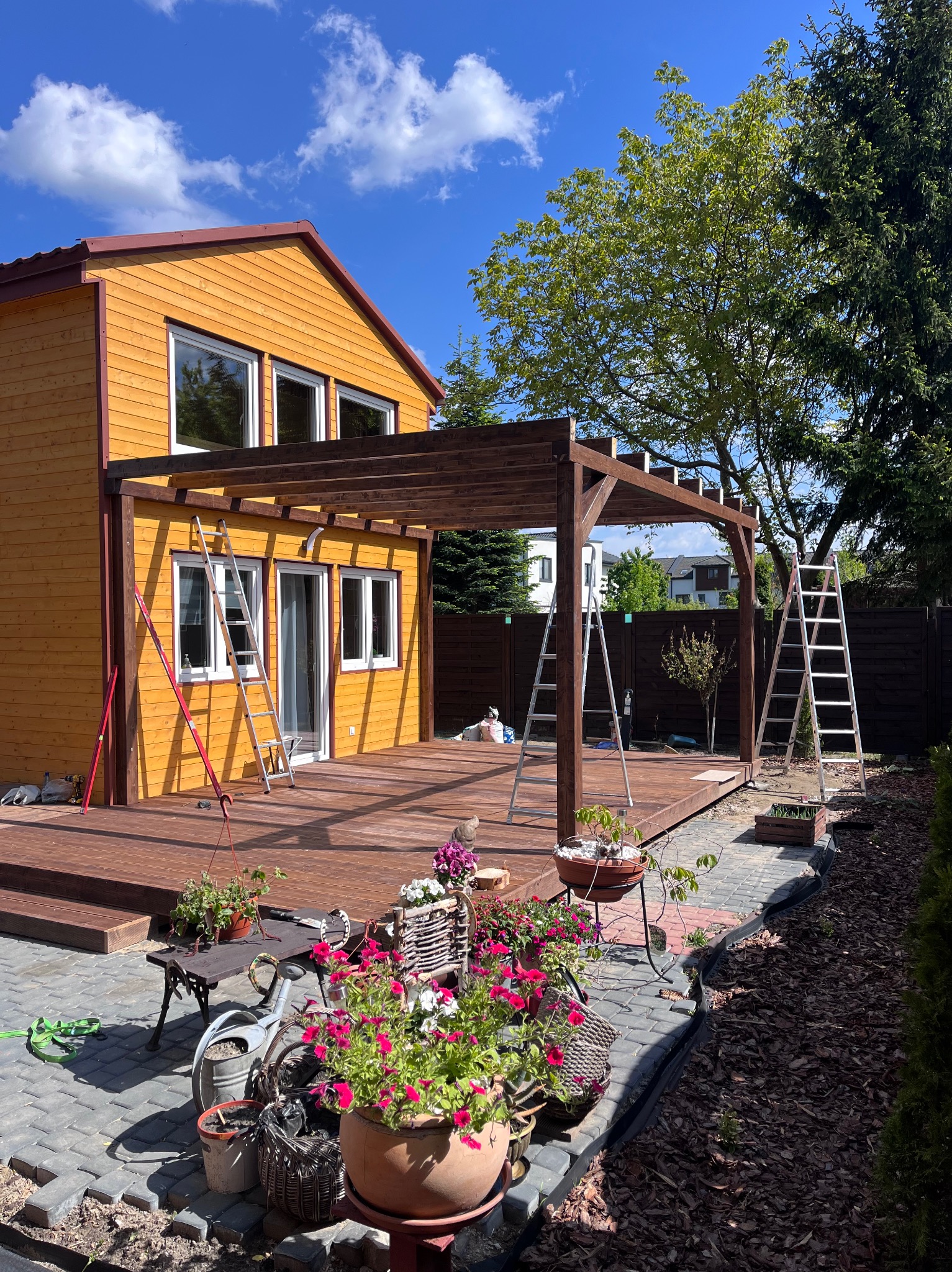 Żółty dom z drewnianą pergolą w trakcie montażu, widoczne drabiny i kwiaty w donicach. Ujęcie w słoneczny dzień z niebieskim niebem.