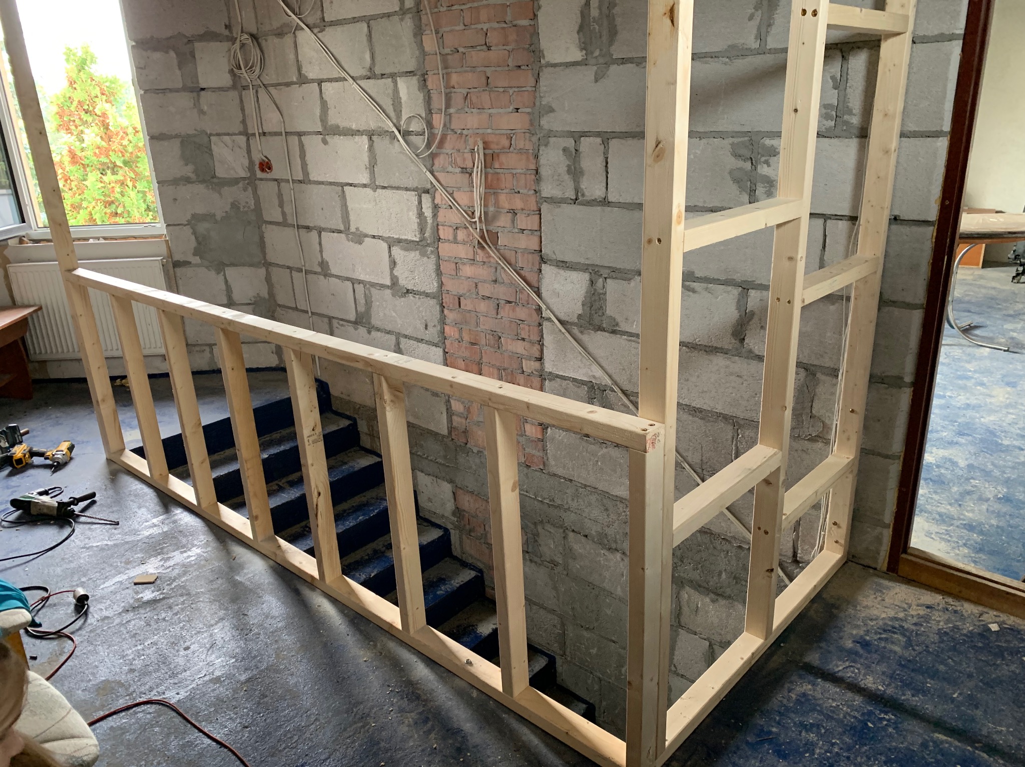 Konstrukcja drewniana schodów w surowym wnętrzu. Widoczne narzędzia i przewody, ściany z cegły i bloczków. Etap budowy balustrady schodowej.