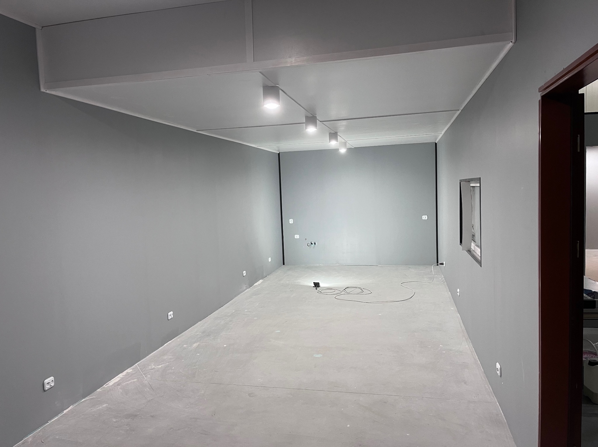 Szare, puste wnętrze po remoncie z nowym oświetleniem punktowym na suficie podwieszanym i widocznymi gniazdkami elektrycznymi na ścianach.