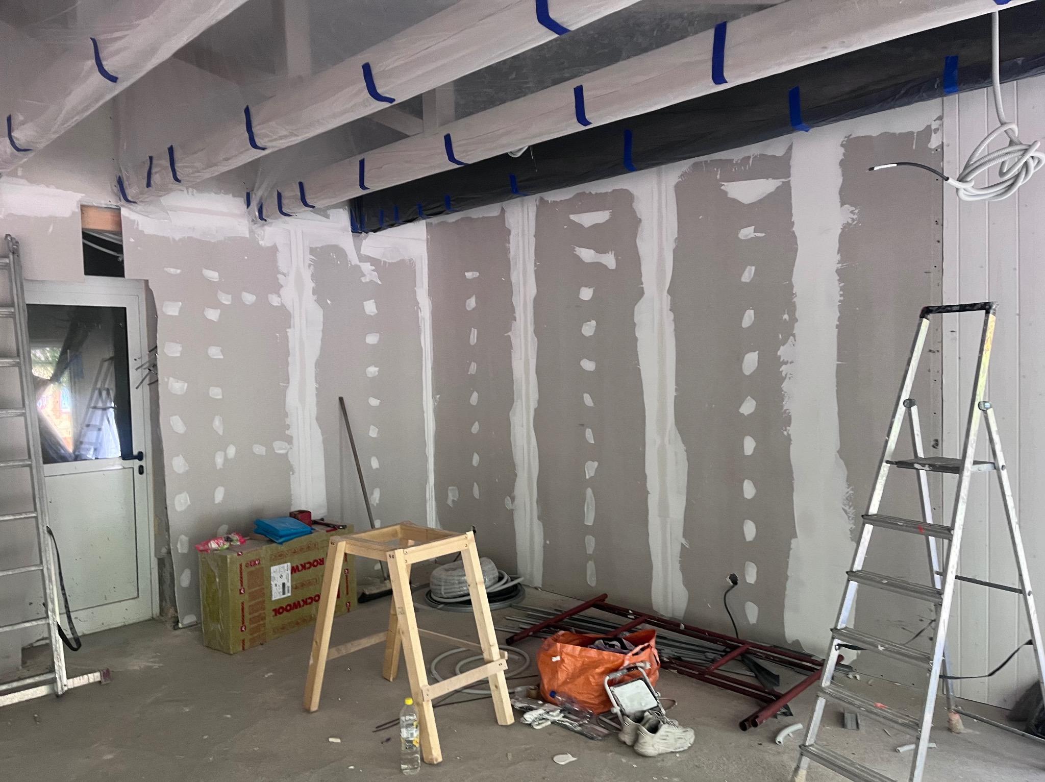 Wnętrze w trakcie wykańczania: ściany z płyt gipsowo-kartonowych, częściowo zaszpachlowane, zabezpieczone folią elementy sufitu, drabiny, narzędzia i materiały budowlane.