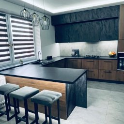 Meble KAVARI - Nowoczesna kuchnia z wyspą: czarne blaty, drewniane fronty szafek, trzy szare hokery. Nad wyspą wiszą trzy metalowe lampy. Okno z żaluzjami w tle.