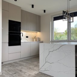 Meble KAVARI - Jasna kuchnia z wyspą i blatem imitującym marmur. Czarne AGD w zabudowie, drewnopodobna podłoga, nowoczesna lampa. Minimalistyczny design, naturalne światło.