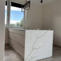 Meble KAVARI - Beżowa wyspa kuchenna z blatem imitującym marmur, oświetlona nowoczesną lampą wiszącą. Widok na okno z roletami i fragment podłogi z paneli.