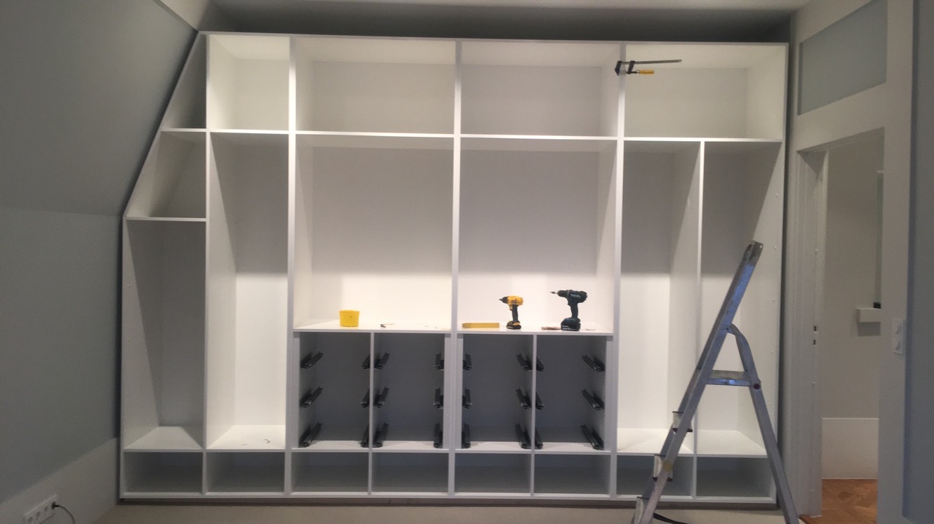 Montaż białej szafy wnękowej z półkami, widoczne narzędzia: wiertarki, ścisk stolarski i drabina aluminiowa.