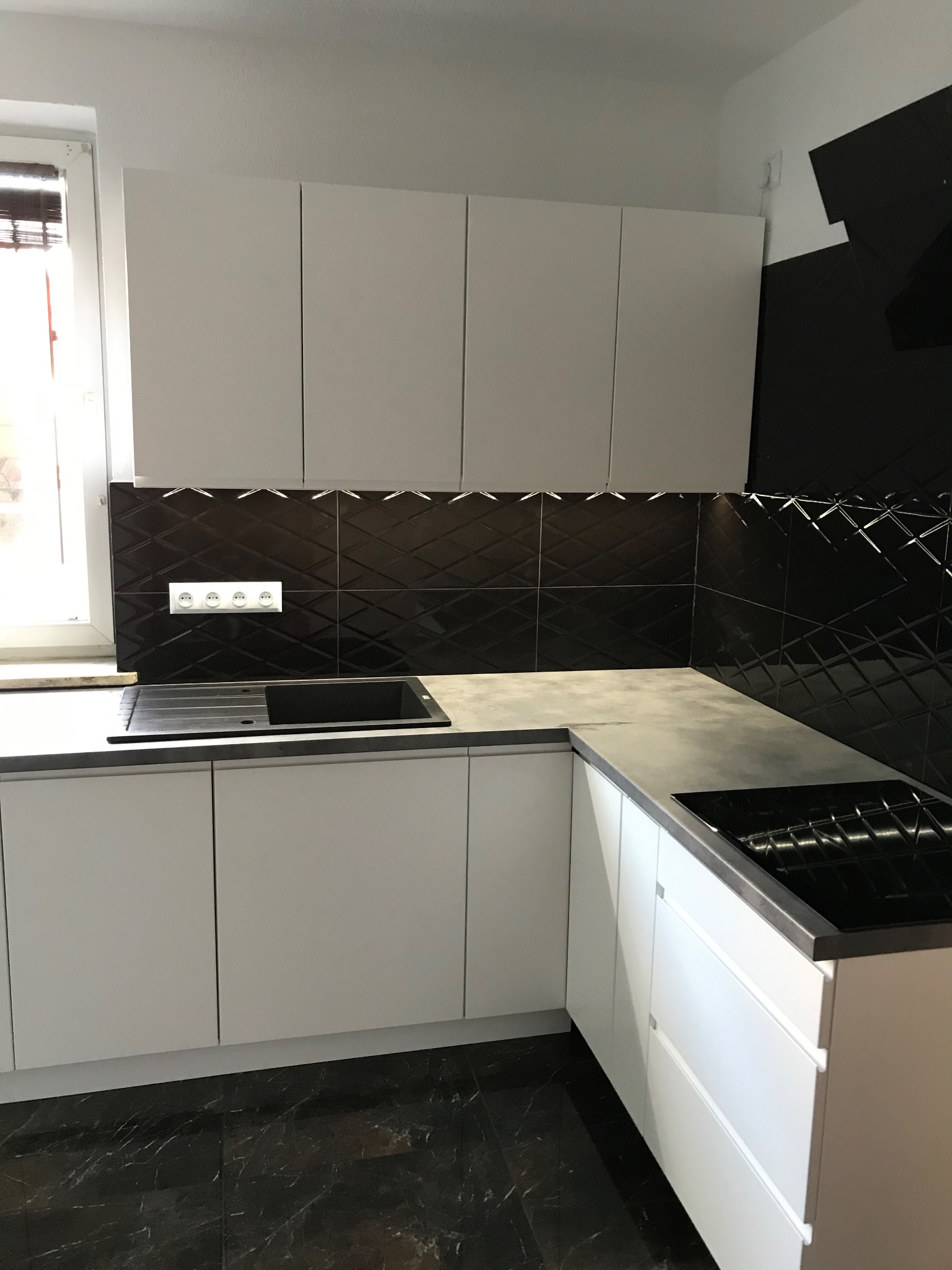 Nowoczesna kuchnia z białymi, gładkimi frontami szafek, czarną, błyszczącą mozaiką na ścianie nad blatem i czarnym zlewozmywakiem oraz płytą indukcyjną. Marmurowe płytki podłogowe.