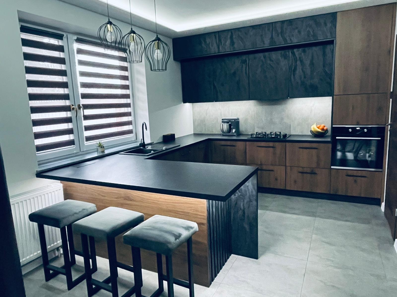 Nowoczesna kuchnia z wyspą: czarne blaty, drewniane fronty szafek, trzy szare hokery. Nad wyspą wiszą trzy metalowe lampy. Okno z żaluzjami w tle.