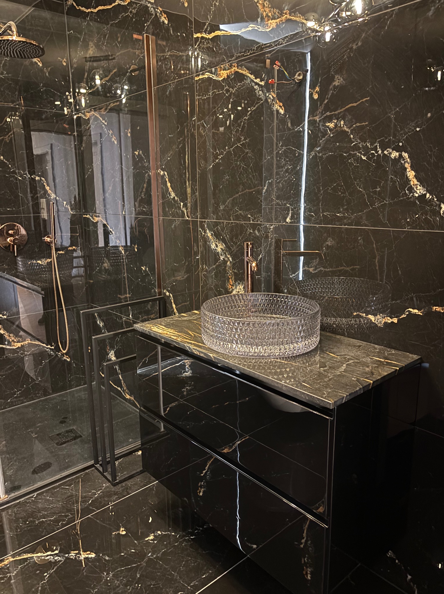 Czarna, marmurkowa łazienka z kryształową umywalką i złotą armaturą. Widoczna kabina prysznicowa z deszczownicą. Elegancki i nowoczesny design.