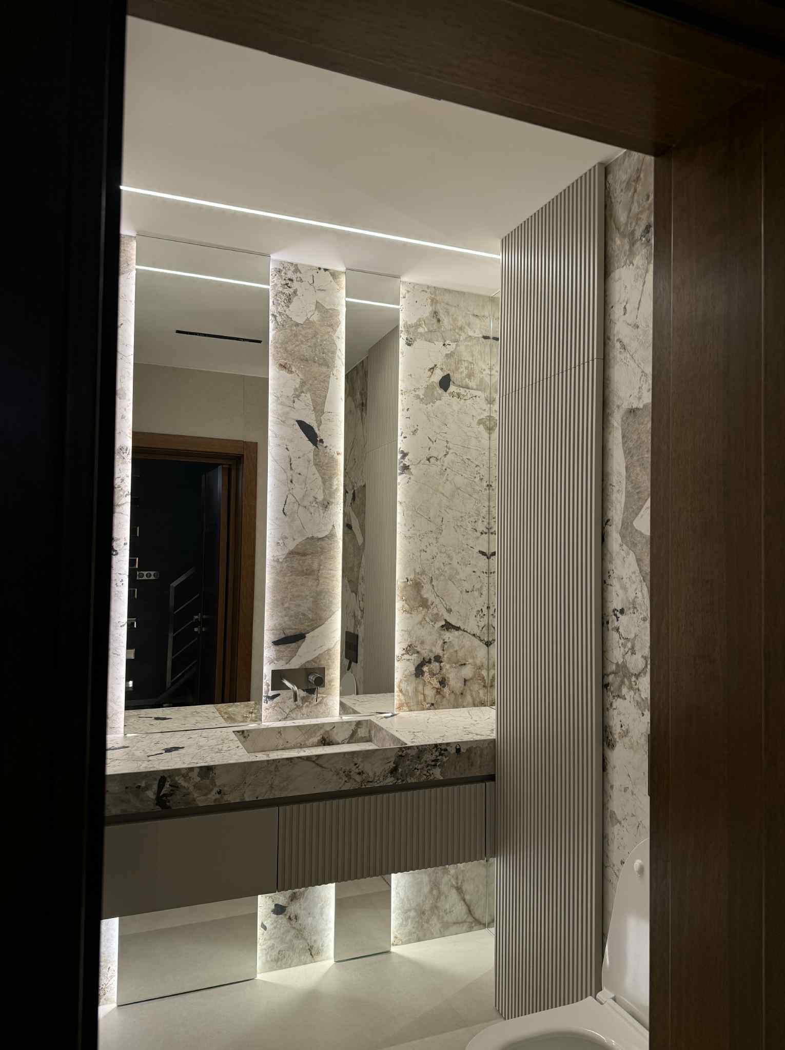 Nowoczesna łazienka z marmurową umywalką i podświetlanymi kolumnami. Widoczne drzwi i fragment toalety. Minimalistyczny design z elementami dekoracyjnymi.