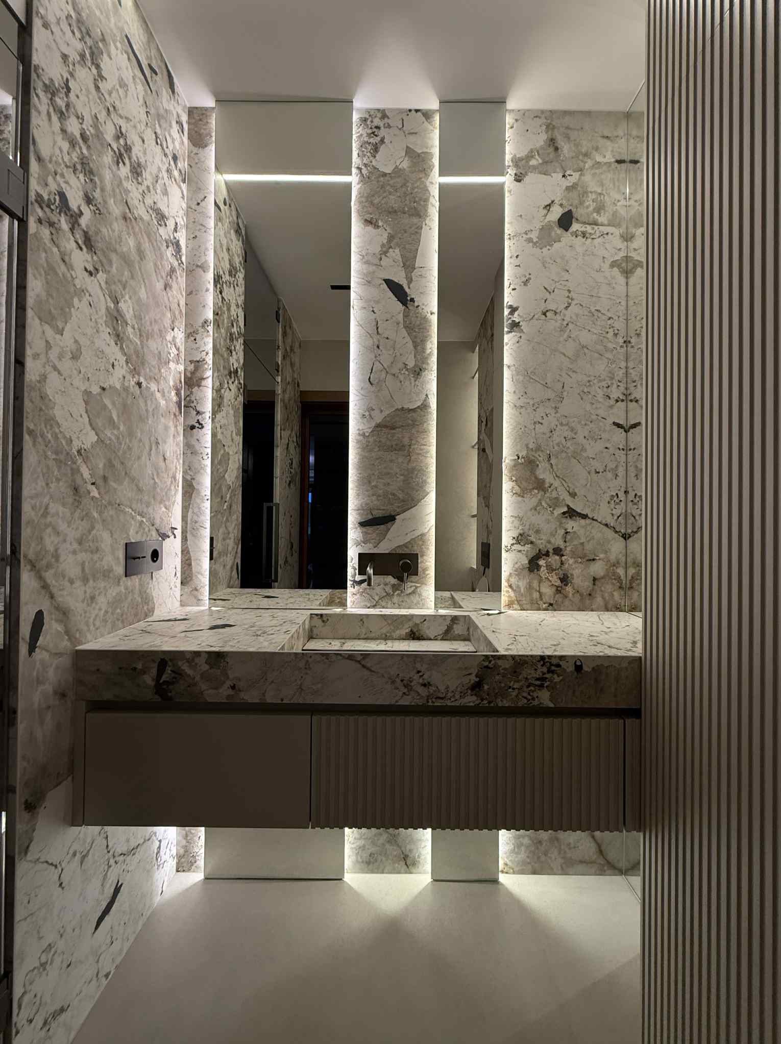 Nowoczesna łazienka z marmurową zabudową umywalki, podświetlanymi lustrami i dekoracyjnymi panelami ściennymi. Minimalistyczny design z subtelnym oświetleniem.