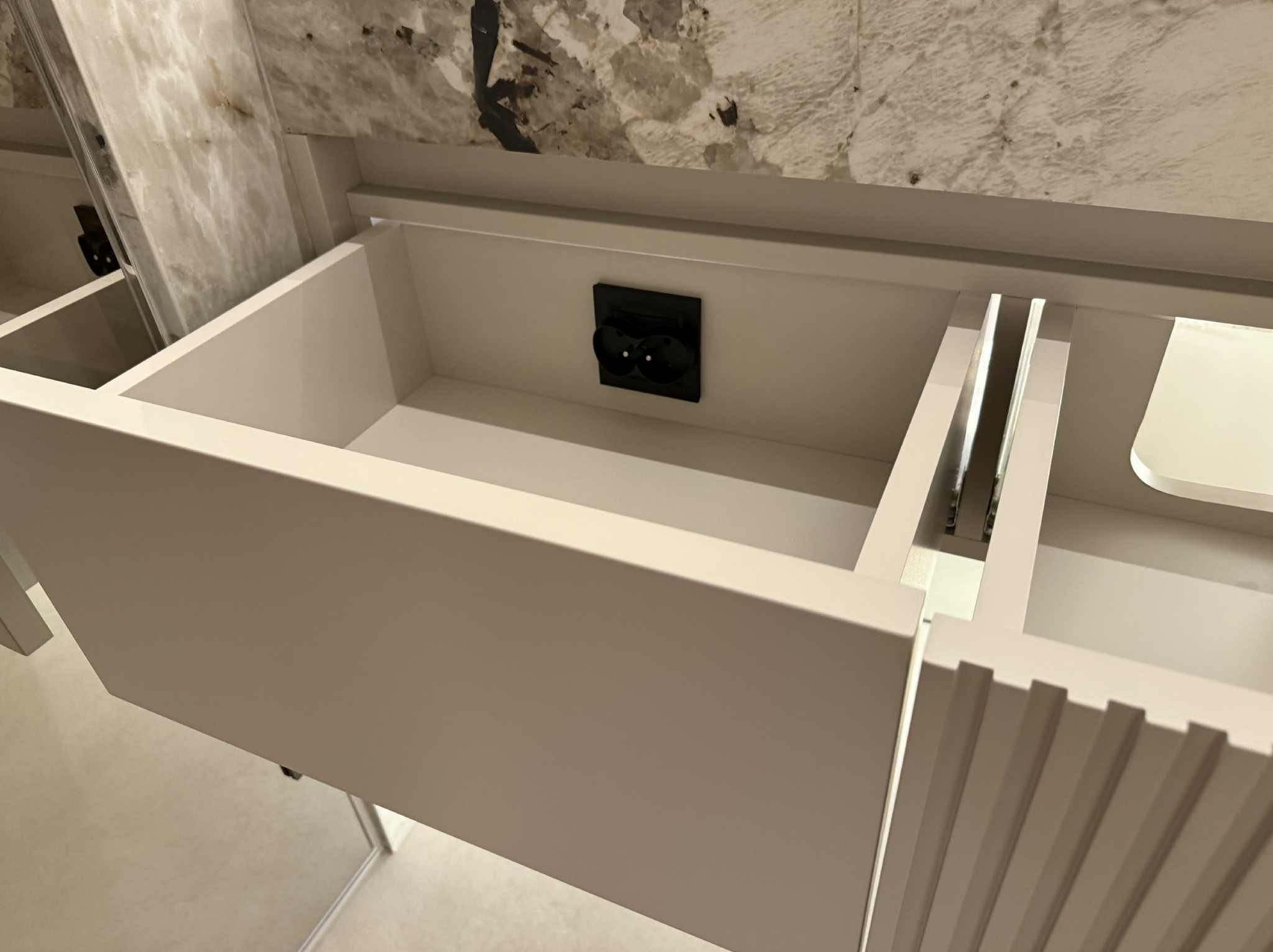Otwarta szuflada w jasnej szafce łazienkowej z marmurowym blatem i gniazdkiem elektrycznym wewnątrz, ukazująca detale wykończenia i funkcjonalność.