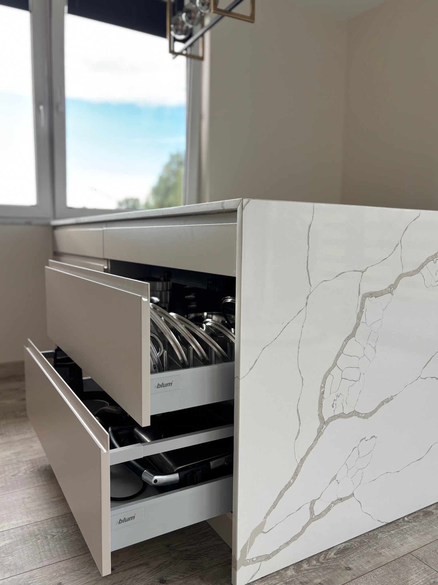 Beżowa szafka kuchenna z blatem imitującym marmur, otwarte szuflady z systemem Blum, widoczne garnki i akcesoria kuchenne. Ujęcie z bliska, naturalne światło.