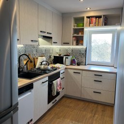 Alder Project - Jasna kuchnia z drewnianymi frontami i czarnymi uchwytami. Nad blatem dekoracyjne płytki, a nad oknem półki z książkami. Na lodówce magnes w kształcie serca.