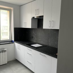 Alder Project - Biała kuchnia z czarnym blatem i marmurowym panelem nad blatem. Minimalistyczne szafki z podłużnymi uchwytami. Widok na okno i grzejnik.