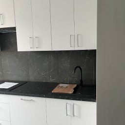 Alder Project - Białe szafki kuchenne z czarnym blatem i marmurowym backsplashiem. Minimalistyczny design, srebrne uchwyty, czarna bateria. Na blacie deska do krojenia i dokumenty.