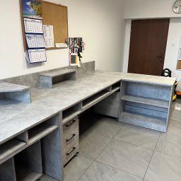 Alder Project - Szary, narożny blat z szafkami i szufladami na kółkach, z kamiennym wzorem, w jasnym pomieszczeniu z płytkami podłogowymi i kalendarzem na ścianie.