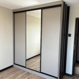 Alder Project - Jasna szafa wnękowa z przesuwnymi drzwiami i lustrem, wykończona czarnymi detalami, na tle drewnianej podłogi. Widoczne pionowe panele dekoracyjne po prawej stronie.