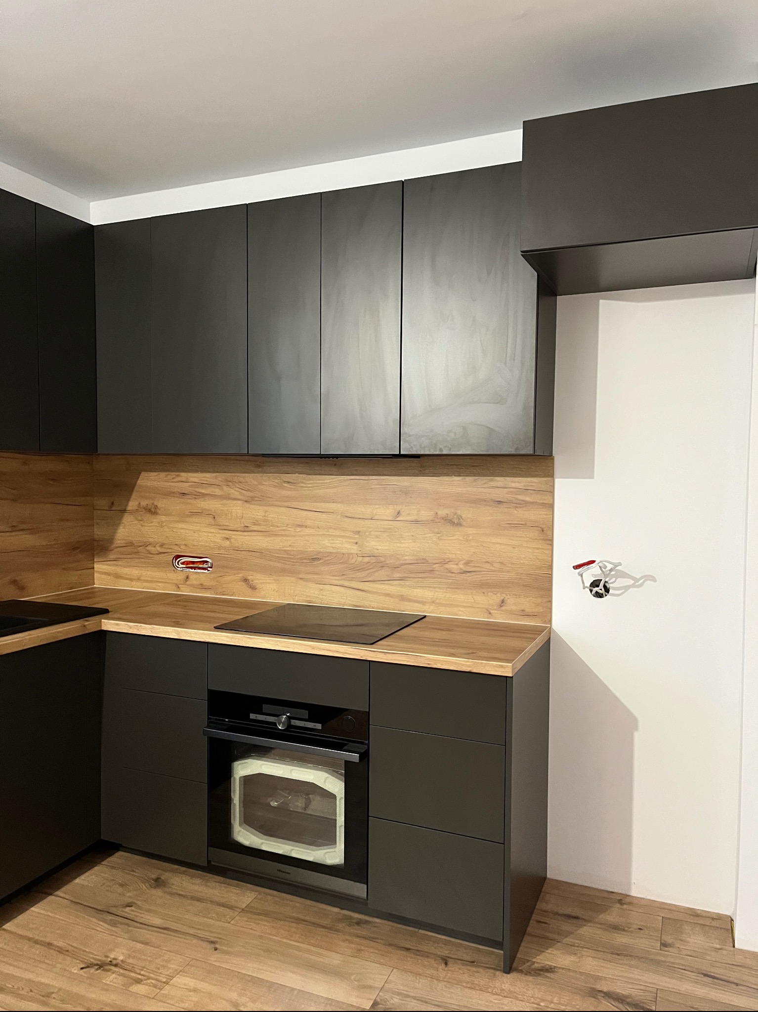 Nowoczesna kuchnia na wymiar z czarnymi frontami i drewnianym blatem. Widoczna płyta indukcyjna i piekarnik. Na ścianie gniazdko elektryczne i zawór.