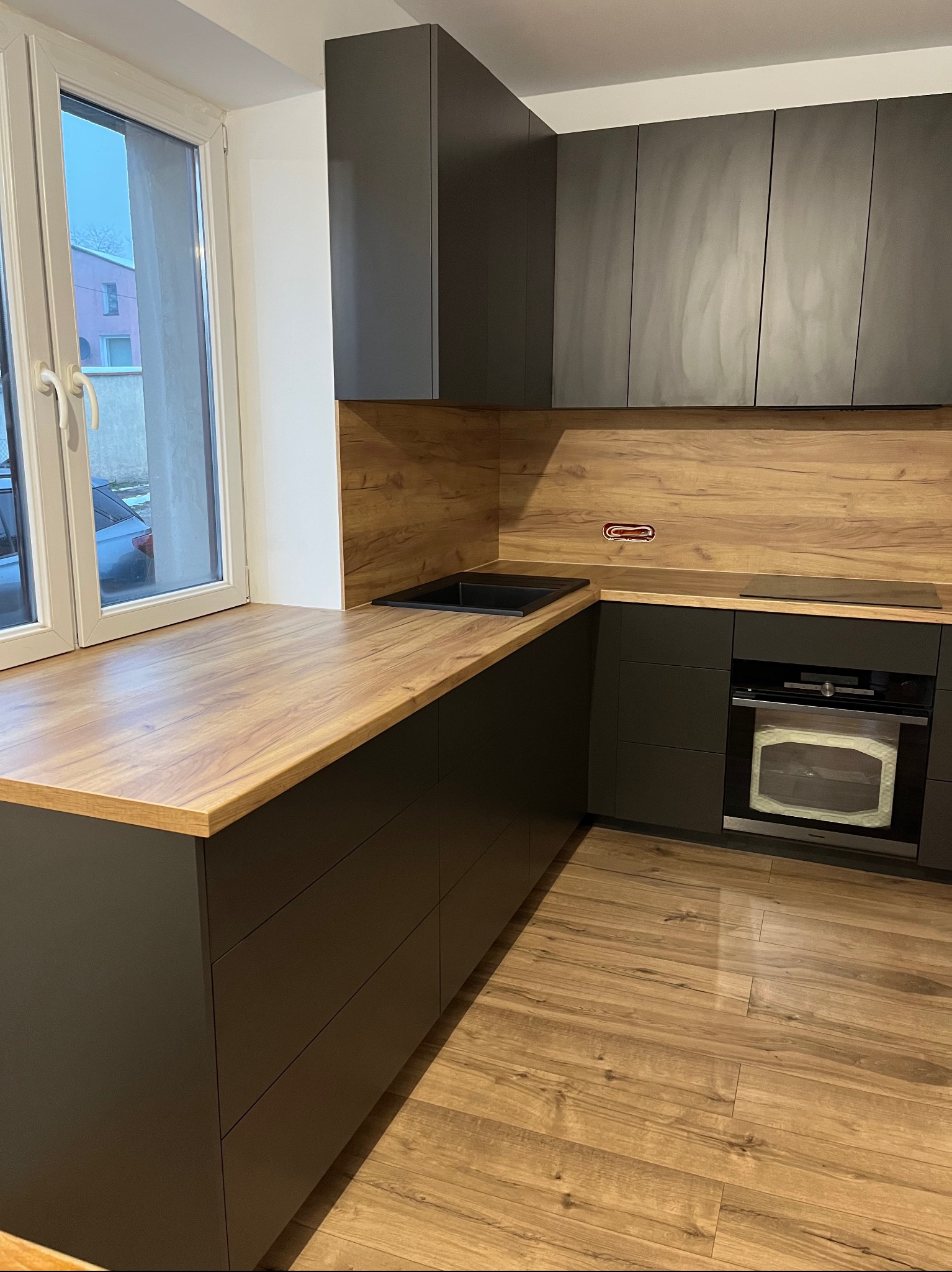 Nowoczesna kuchnia z ciemnymi szafkami i drewnianym blatem przy oknie. Widoczny piekarnik i płyta indukcyjna. Podłoga drewniana, ciepłe oświetlenie.