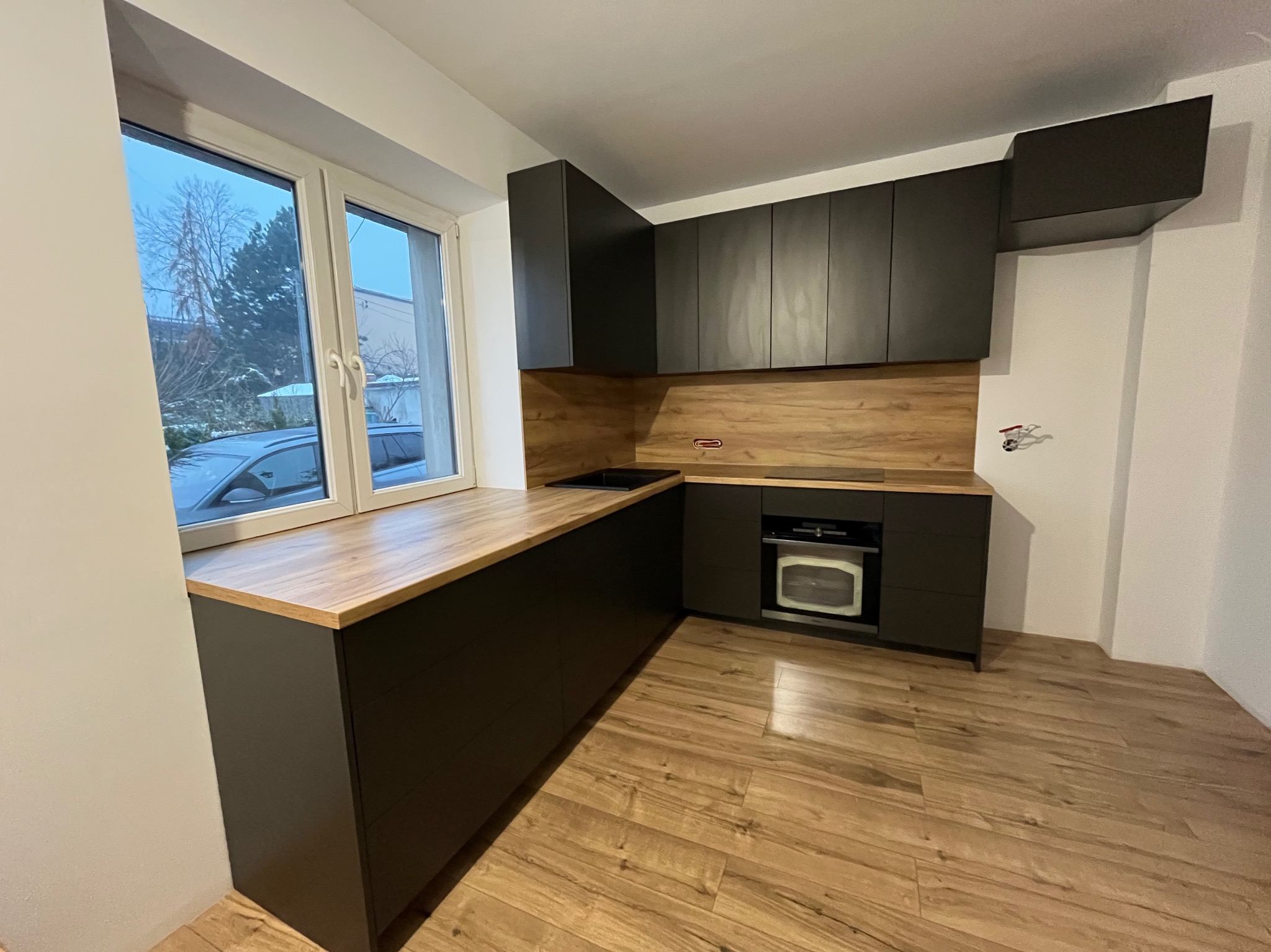 Nowoczesna kuchnia z czarnymi szafkami i drewnianym blatem przy oknie. Widoczny piekarnik i drewniana podłoga. Minimalistyczny design.