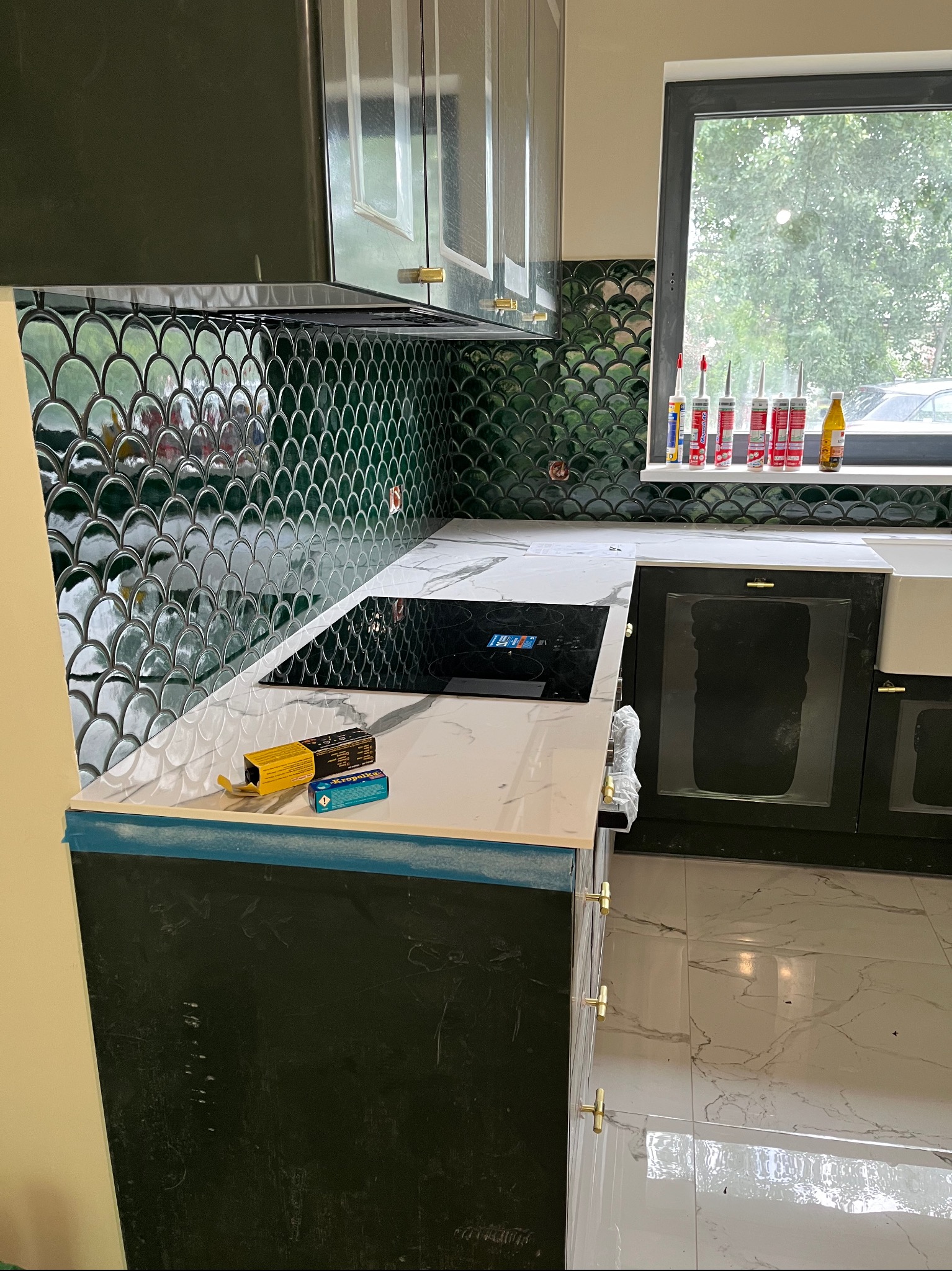 Montaż kuchni: zielona mozaika na ścianie, biały blat z płytą indukcyjną, ciemne szafki z złotymi uchwytami. Widoczne narzędzia i silikony.