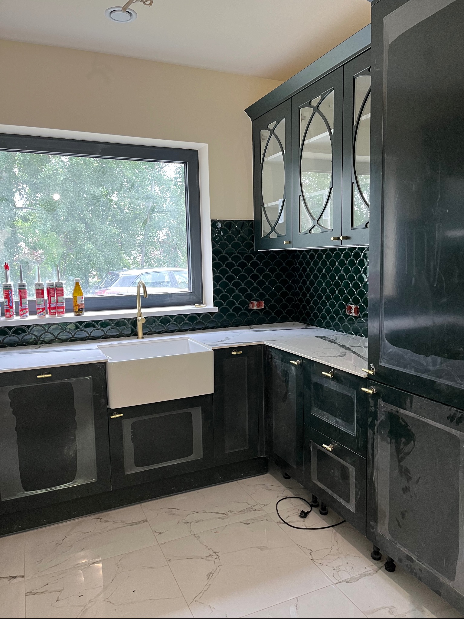 Nowoczesna kuchnia z ciemnozielonymi szafkami, białym blatem i zlewozmywakiem farmerskim. Uwagę przyciągają płytki w kształcie rybiej łuski i złota armatura.