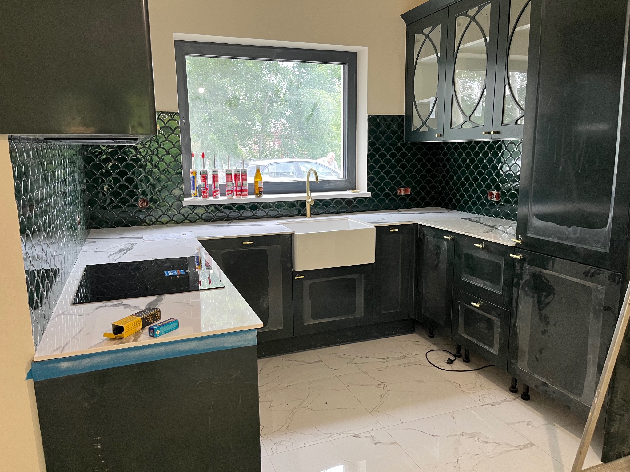 Ciemnozielona kuchnia z białym blatem i zlewozmywakiem farmerskim, wykończona mozaiką w kształcie rybiej łuski. Widoczne silikony i narzędzia, sugerujące etap montażu.