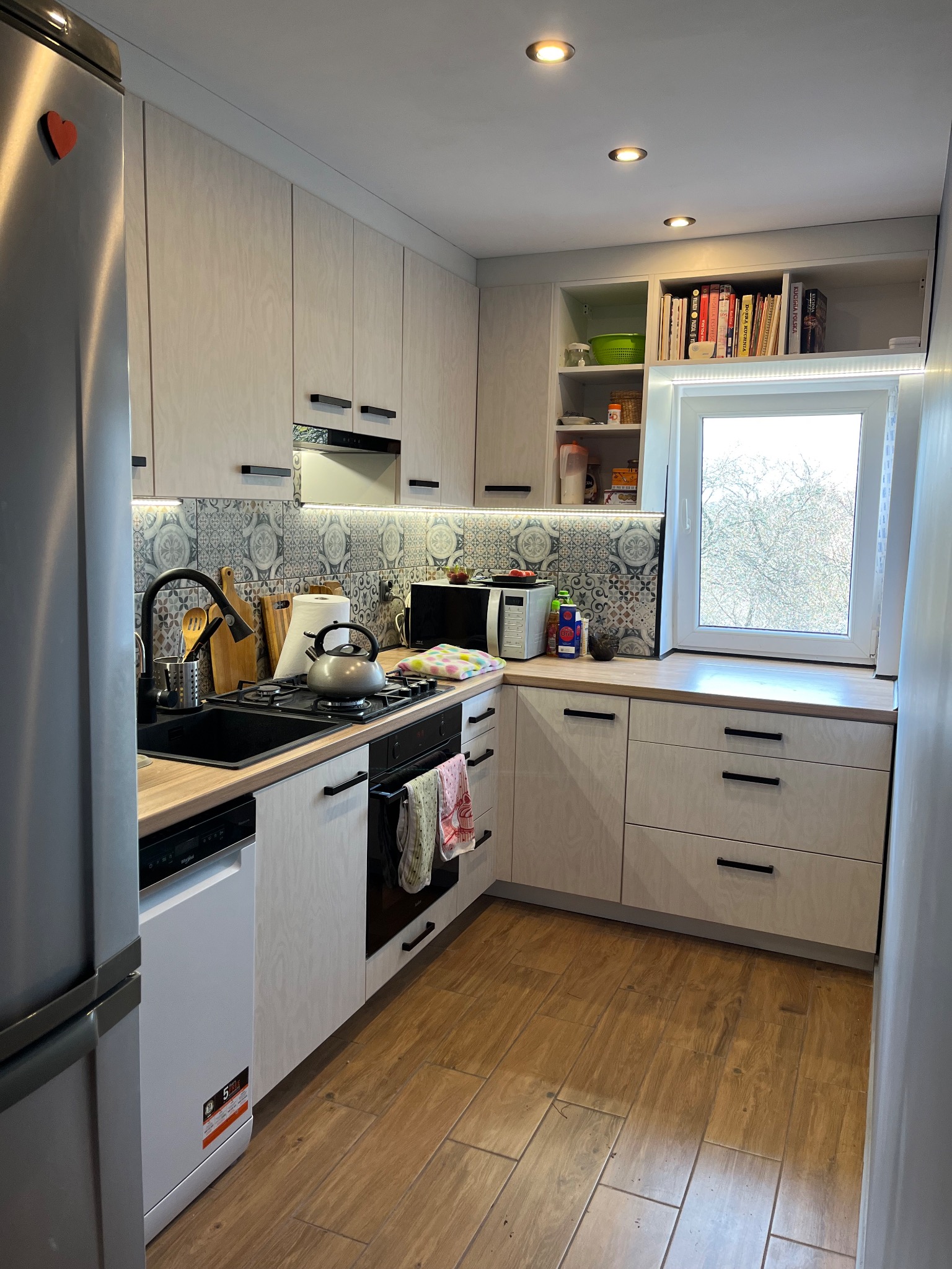 Jasna kuchnia z drewnianymi frontami i czarnymi uchwytami. Nad blatem dekoracyjne płytki, a nad oknem półki z książkami. Na lodówce magnes w kształcie serca.