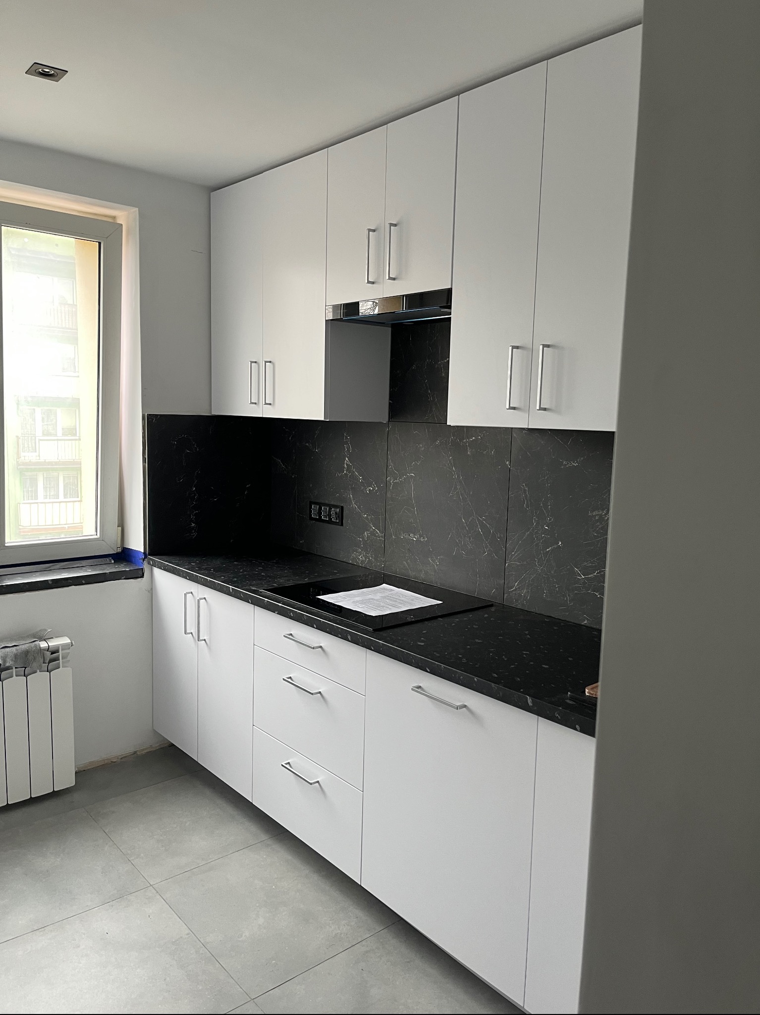 Biała kuchnia z czarnym blatem i marmurowym panelem nad blatem. Minimalistyczne szafki z podłużnymi uchwytami. Widok na okno i grzejnik.