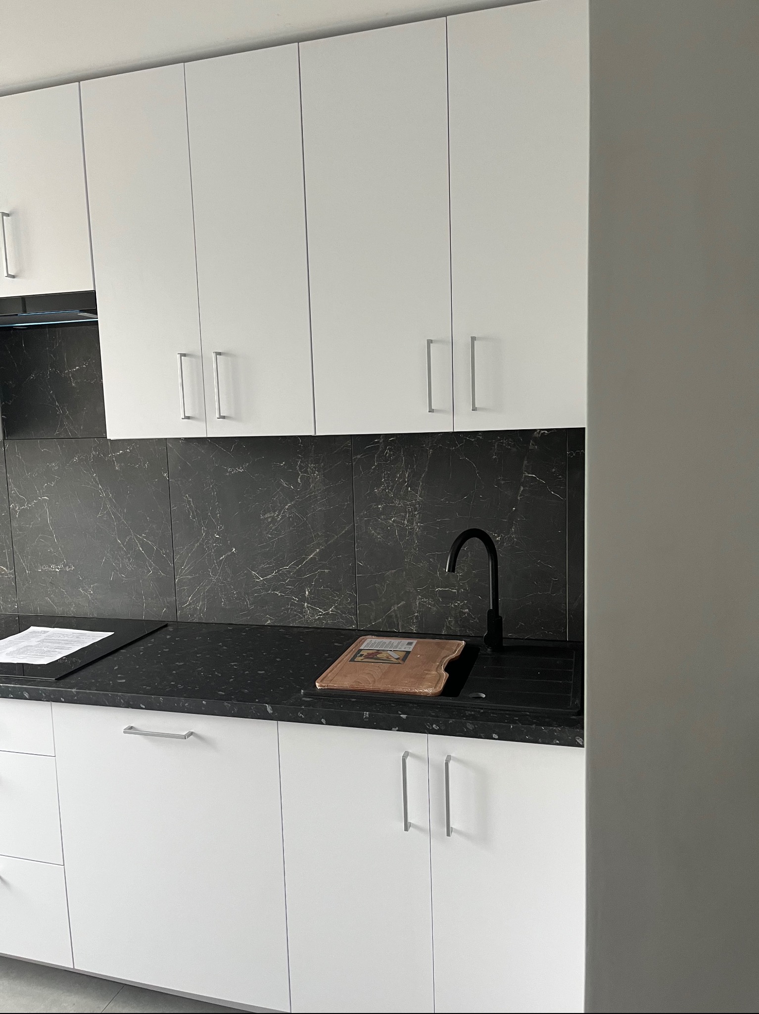 Białe szafki kuchenne z czarnym blatem i marmurowym backsplashiem. Minimalistyczny design, srebrne uchwyty, czarna bateria. Na blacie deska do krojenia i dokumenty.