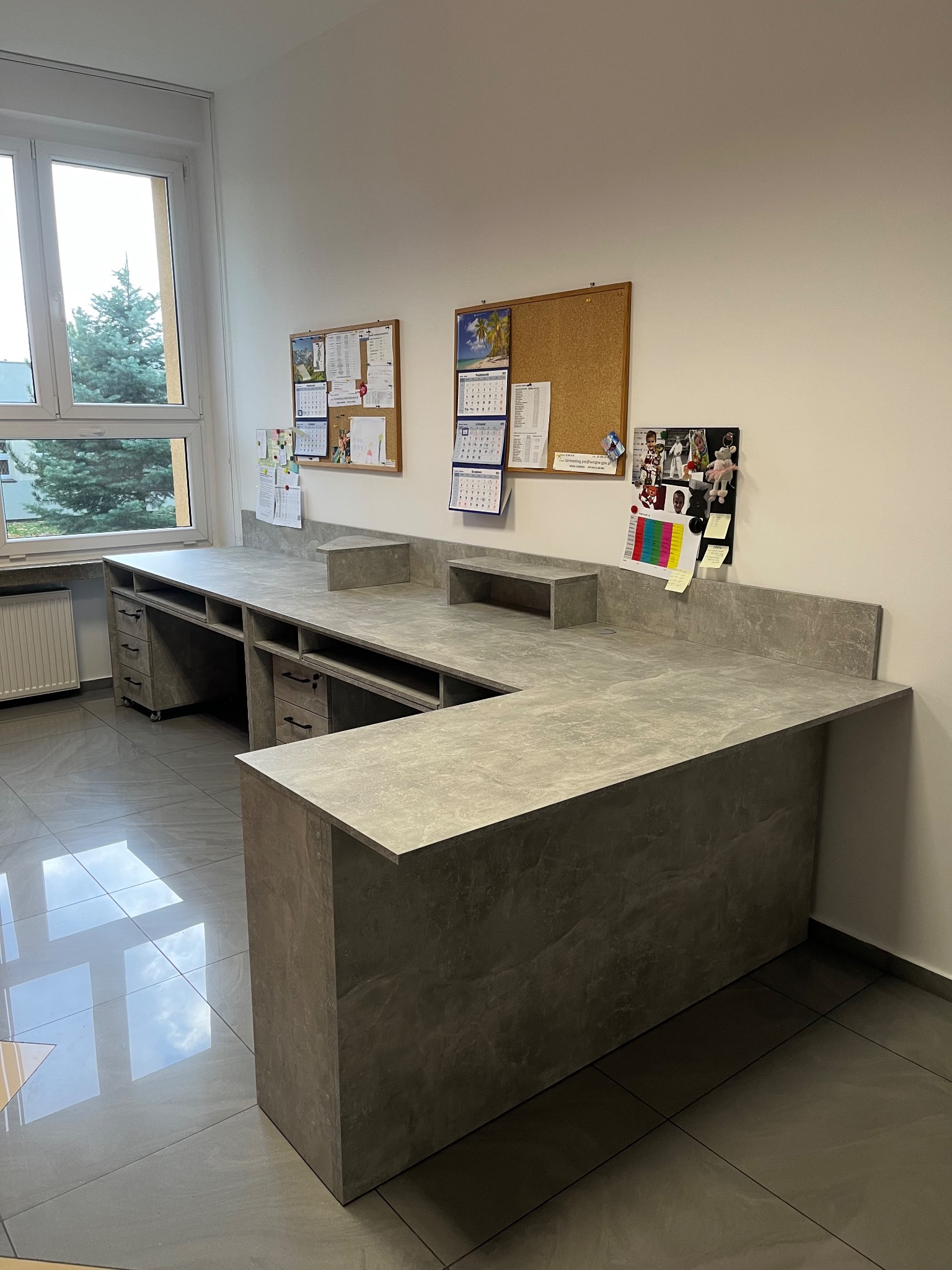Długi, szary blat z szafkami w kształcie litery U, prawdopodobnie w biurze, z kalendarzami i tablicami korkowymi na ścianie. Widok z perspektywy.